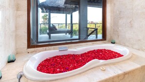 A bathroom with a bathtub filled with red rose petals, a large mirror, and a view of an outdoor seating area. - Villa Lalang Temu (Jimbaran)