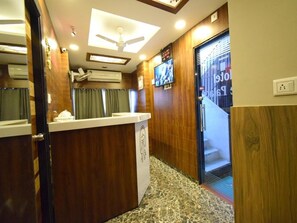 A reception area with a wooden reception desk, a ceiling fan, an air conditioning unit, and a television mounted on the wall. - The Lake Palace (Ahmedabad)