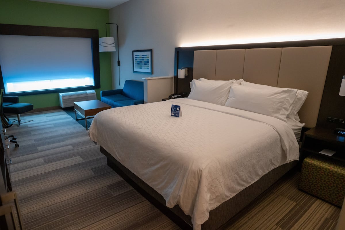 A hotel room with a large bed, a desk, a chair, a window with blinds, and a wall-mounted air conditioner.