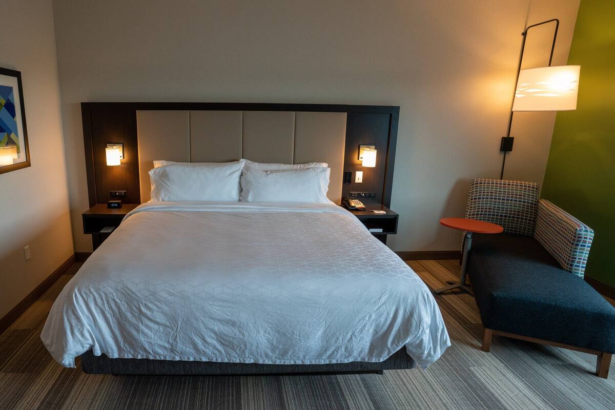 A hotel room with a large bed, two bedside tables with lamps, a small table, a chair, and a framed picture on the wall.