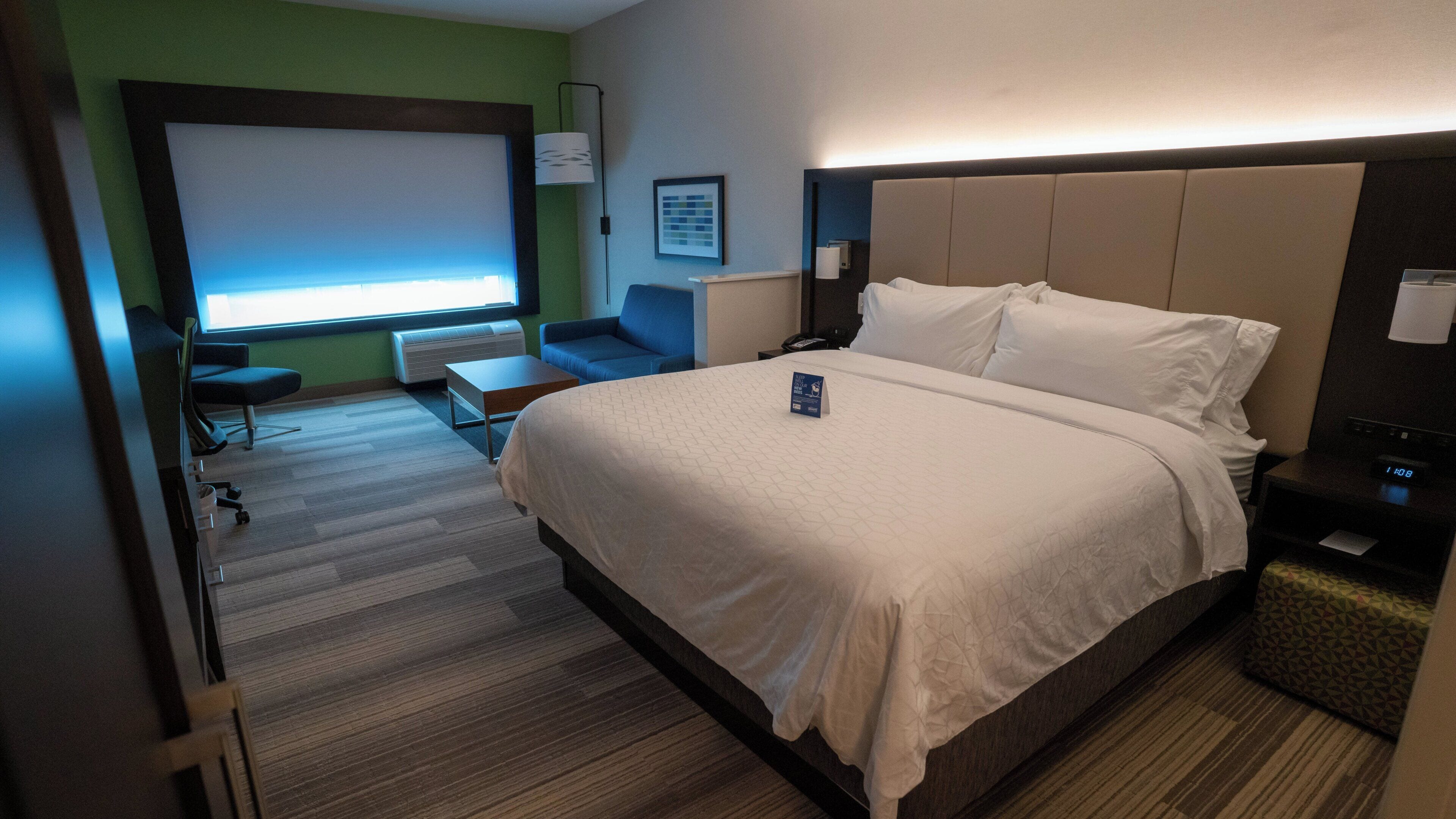 Una habitación de hotel con cama, escritorio, silla, ventana y lámpara de pared.