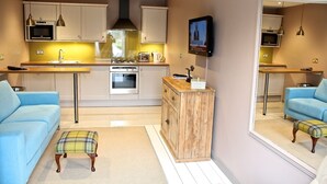 A modern kitchen with a central island, built-in oven, and stainless steel appliances. There is a seating area with a sofa and ottoman, and a television mounted on the wall. - The Apartments Quidhampton Mill (Salisbury)