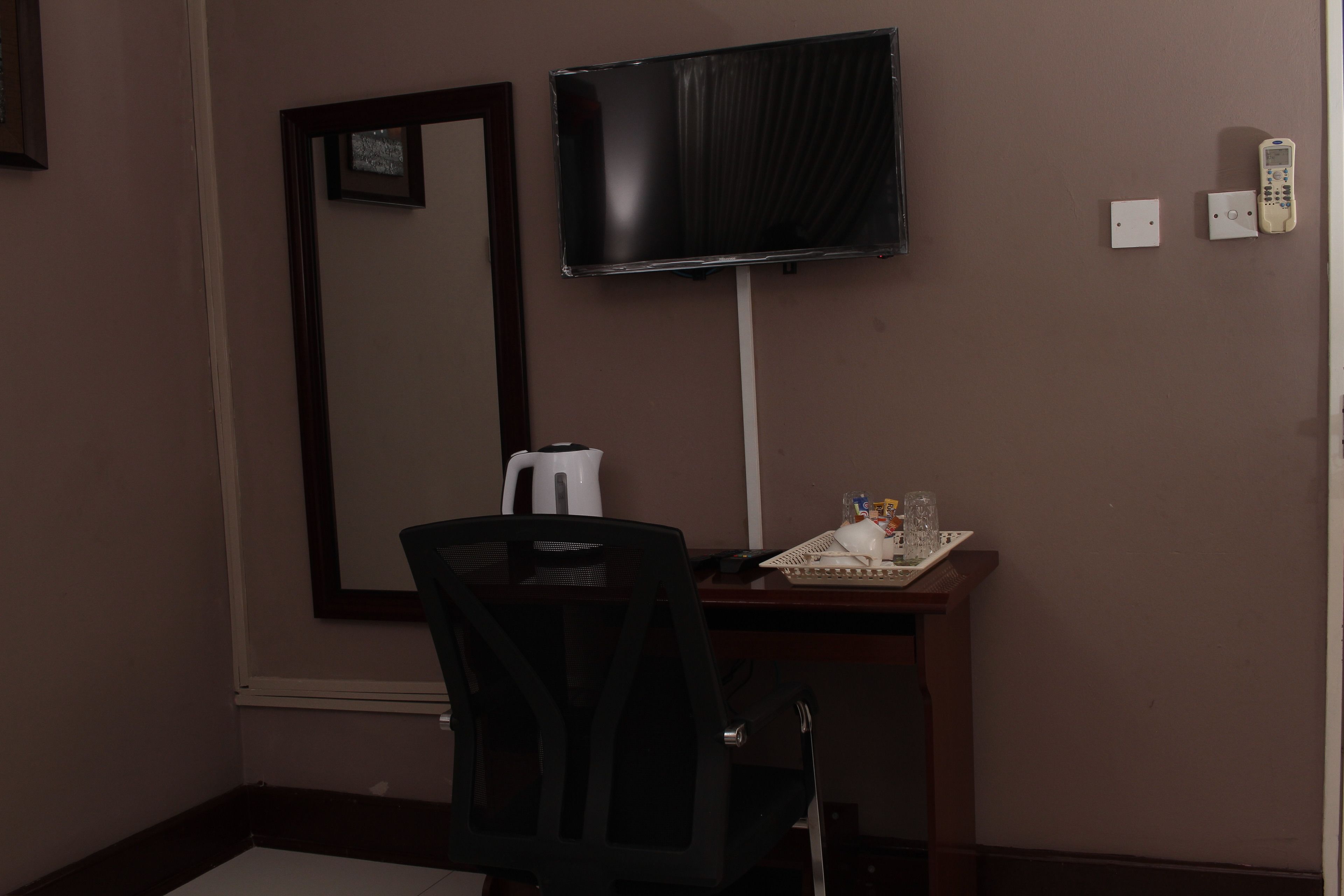Una habitación con escritorio, silla, televisor montado en la pared, teléfono y una mesita con jarra y vasos.