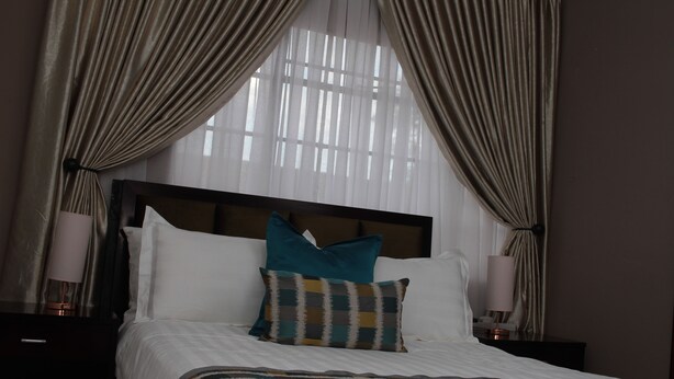 A hotel room with a large bed, two pillows, a headboard, a bedside table with a lamp, and curtains. - Triple Palms Bed and Breakfast (Gaborone)