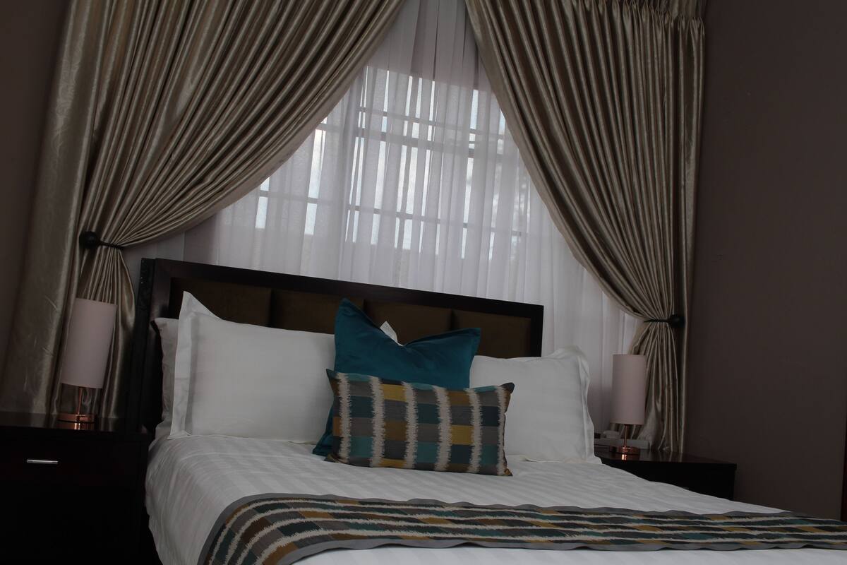 A hotel room with a large bed, two pillows, a headboard, a bedside table with a lamp, and curtains.