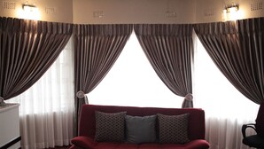 A red sofa with patterned pillows in a room with wooden flooring, large windows with curtains, and a desk with a lamp. - Triple Palms Bed and Breakfast (Gaborone)