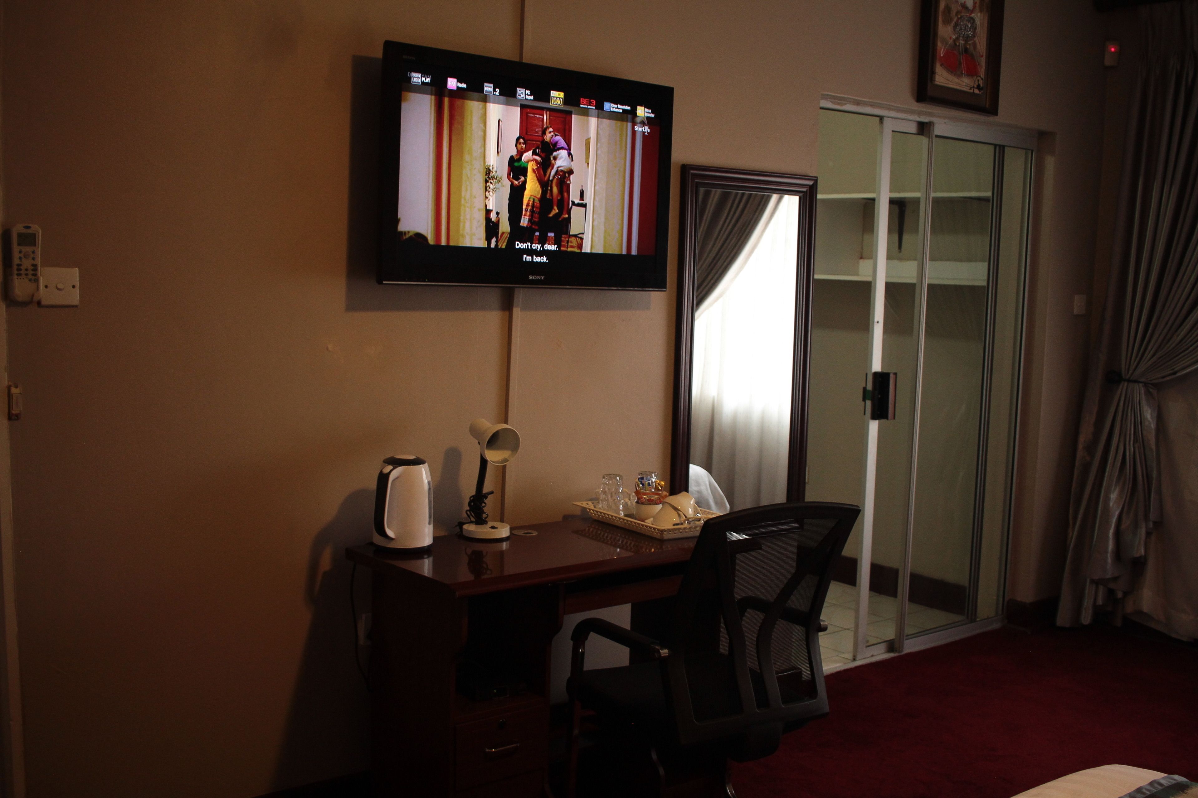 A hotel room with a TV mounted on the wall, a desk with a chair, a coffee maker, a tray with glasses and a plate, and a sliding glass door leading to a balcony.