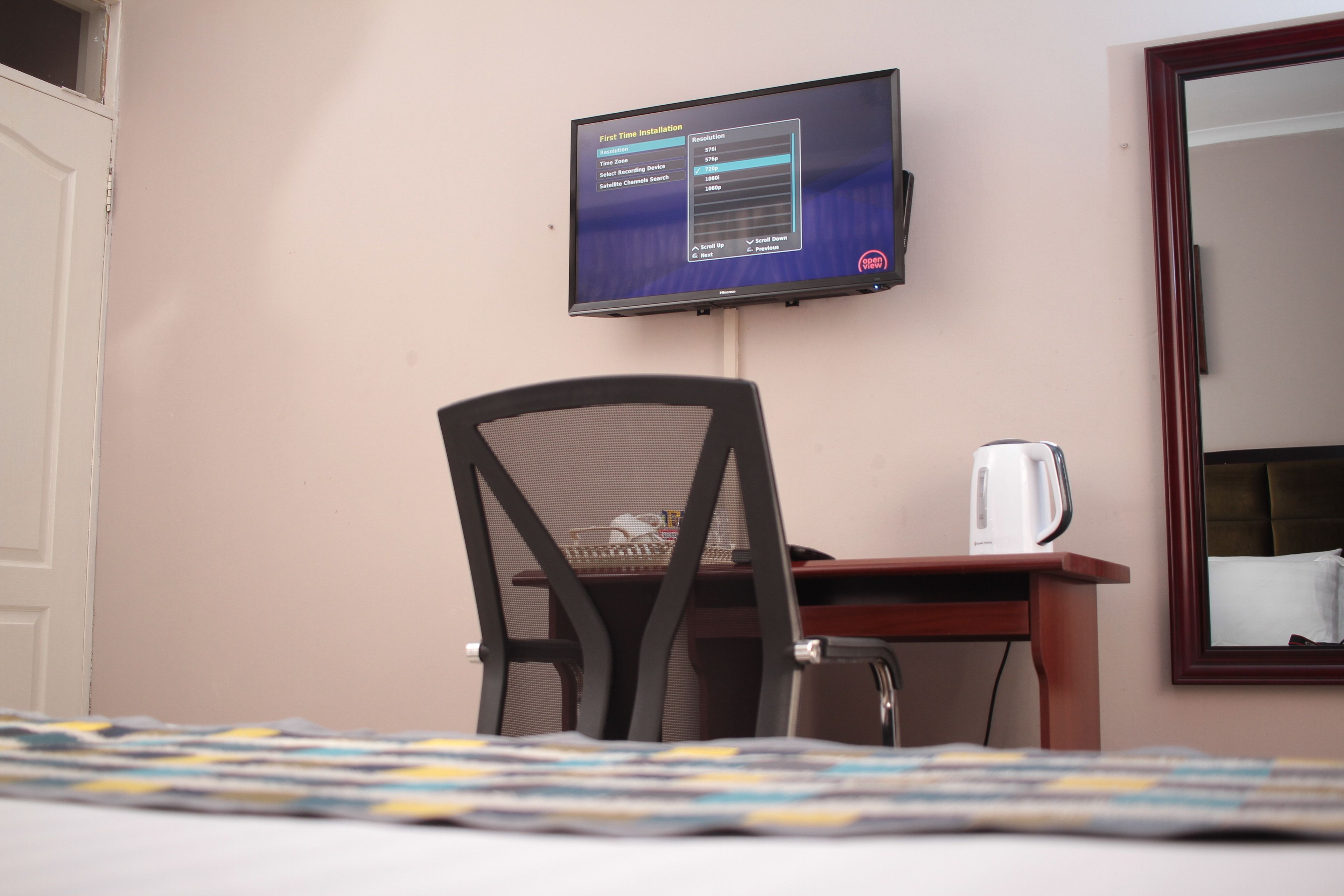 Habitación de hotel con cama, una silla, escritorio con hervidor y televisor mostrando la pantalla de instalación inicial.