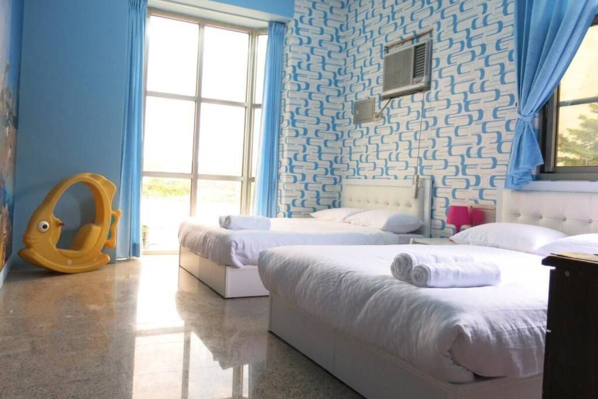 A hotel room with two beds, a window with blue curtains, a patterned wall, and a small air conditioning unit.