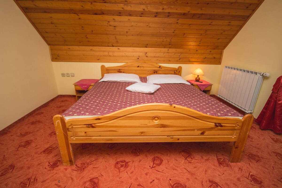 A wooden bed with a headboard, a red patterned bedspread, two pillows, a bedside table with a lamp, and a radiator on the wall.