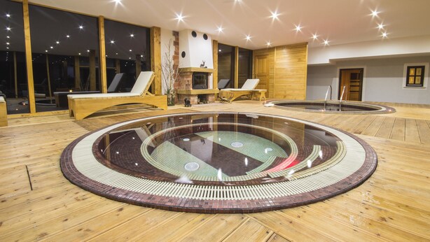 Un área de piscina interior moderna con una piscina circular central, suelo de madera y grandes ventanales.