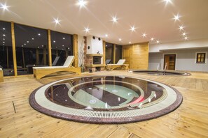 Un área de piscina interior moderna con una piscina circular central, suelo de madera y grandes ventanales.