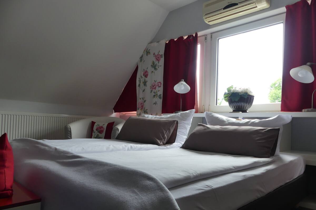 A bedroom with a bed, red curtains, a window with a view, and an air conditioning unit.