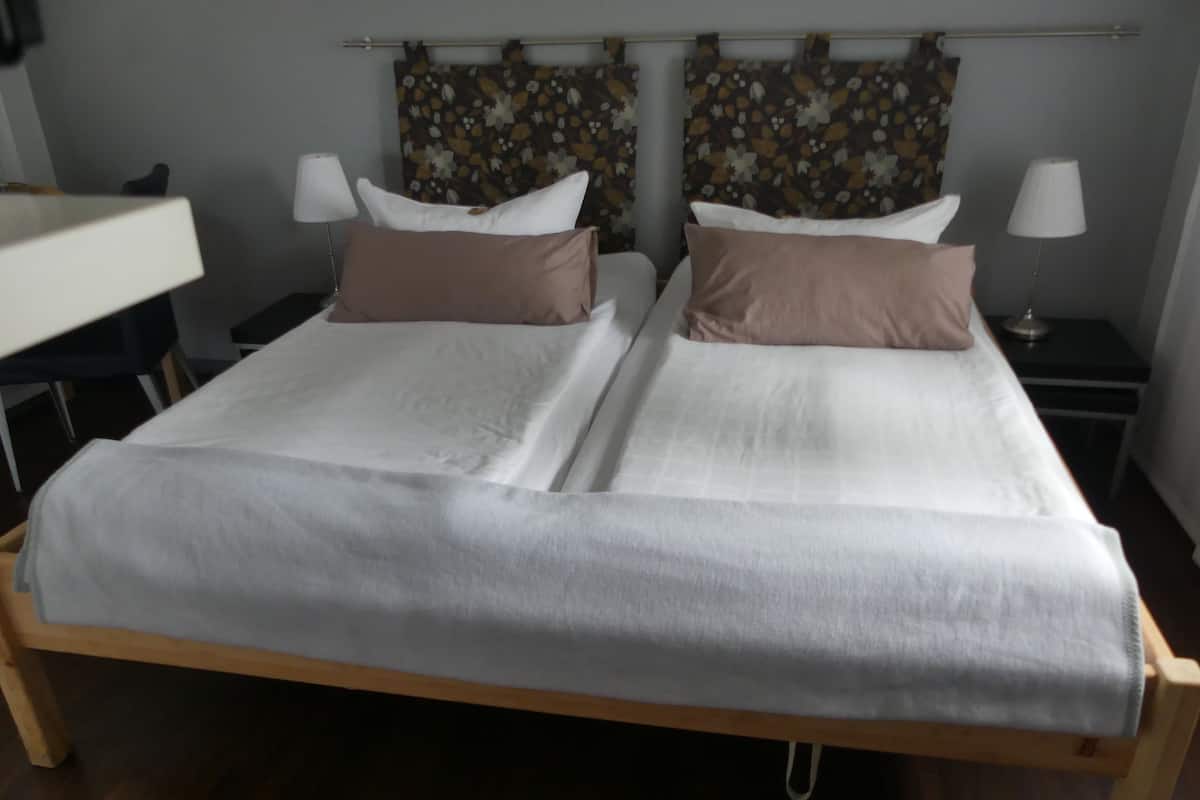 A double bed with white linens and two brown pillows, flanked by nightstands with lamps, in a room with wooden flooring and floral curtains.