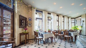 Un comedor con varias mesas y sillas, ventanas amplias con cortinas y piso de baldosas.