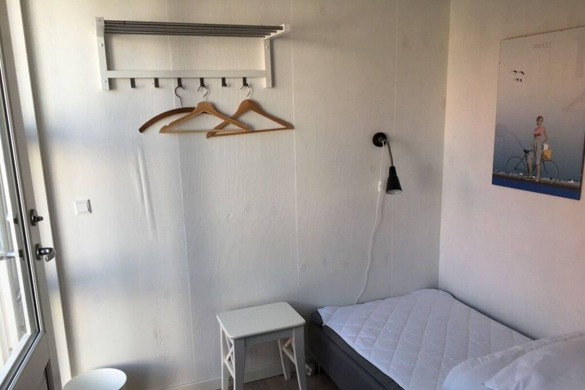 Ein Schlafzimmer mit einem Bett, einem kleinen Tisch, einem Kleiderständer, einer Wandlampe und einem Bild an der Wand.