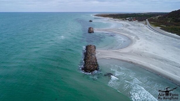 Vue aérienne d’une zone côtière avec une plage de sable, une mer bleue et limpide, et un affleurement rocheux dans l’eau.
