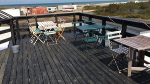 Une terrasse en bois avec des meubles d’extérieur, une vue sur la plage et un bâtiment situé au loin.