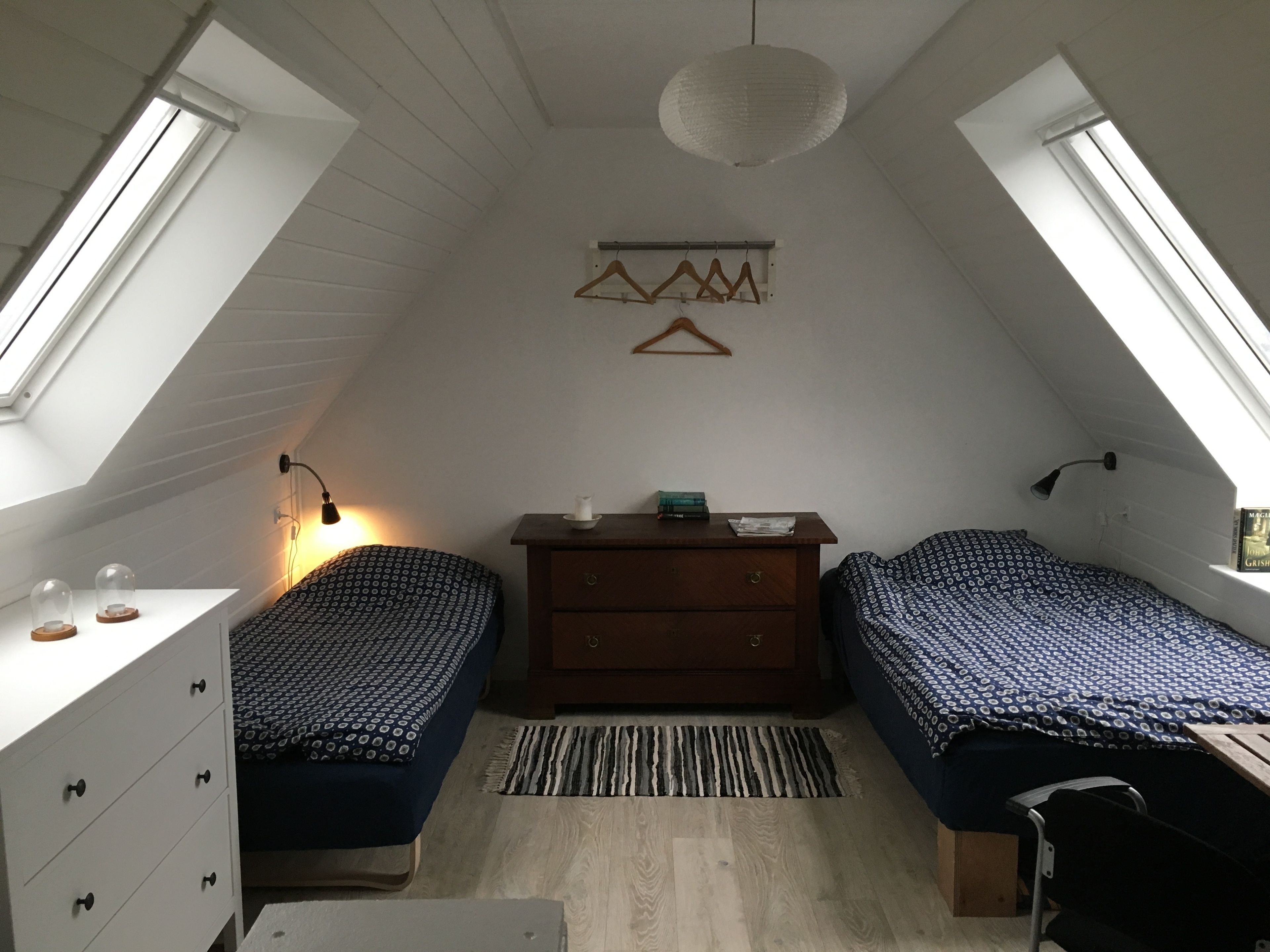 A small, well-lit attic bedroom with two single beds, a dresser, a hanging light fixture, and a window.