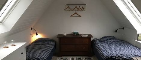 A small, well-lit attic bedroom with two single beds, a dresser, a hanging light fixture, and a window.