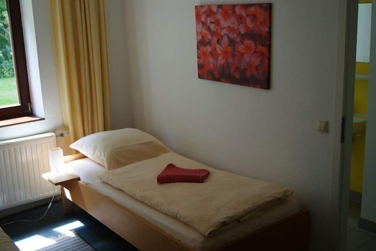 A single bed with a beige mattress, a red blanket, and a pillow. There is a window with curtains, a radiator, and a wall-mounted painting.