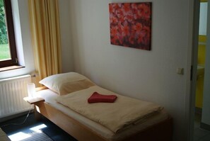 A single bed with a beige mattress, a red blanket, and a pillow. There is a window with curtains, a radiator, and a wall-mounted painting. - Zimmerfrei (Wuppertal)
