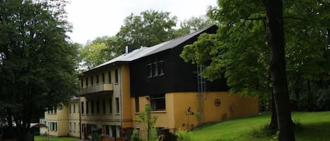 A two-story building with a yellow facade and a black roof, surrounded by greenery and trees.