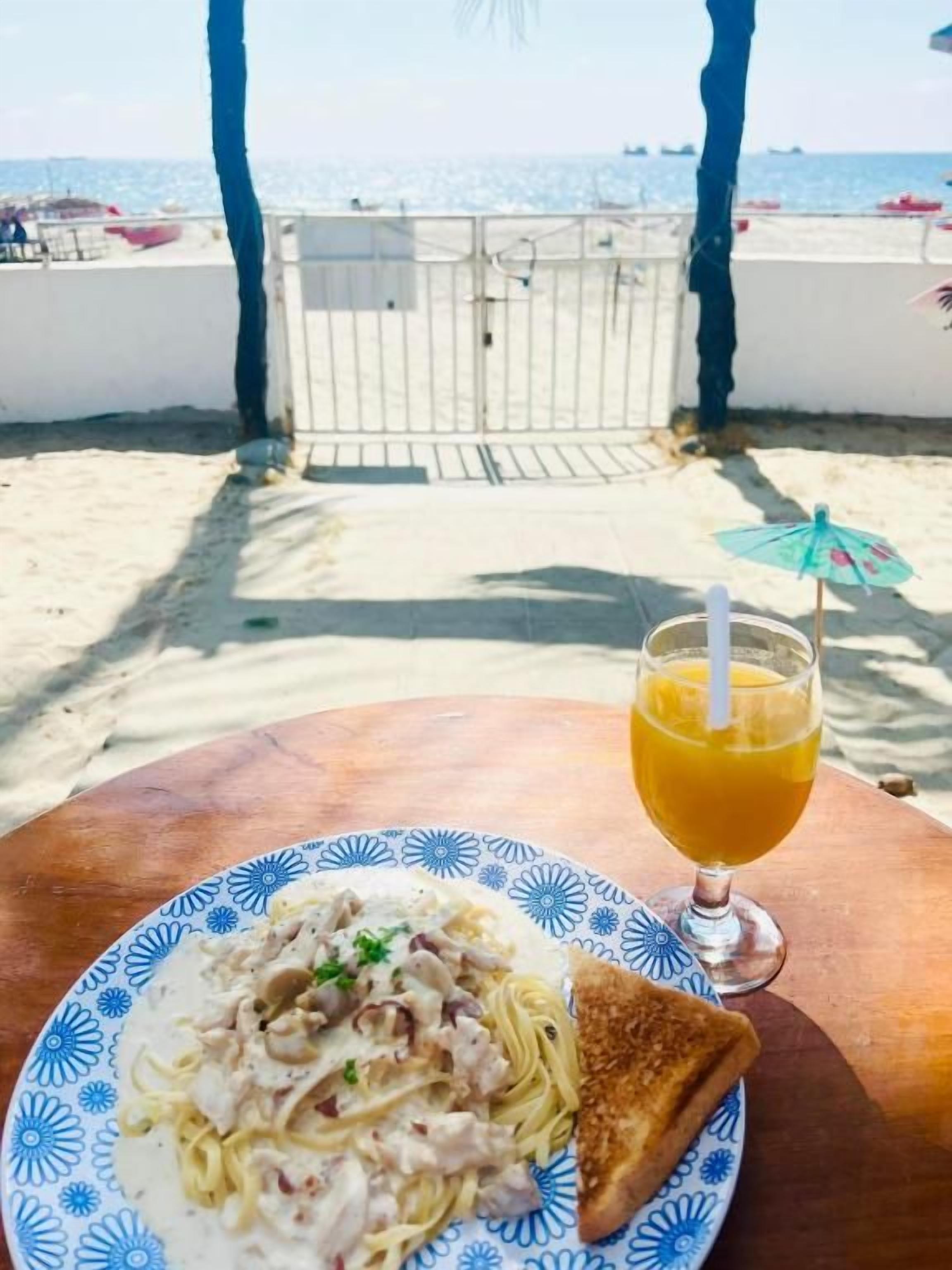 A table with a plate of food and a drink, a beach in the background, and a gate leading to the beach.
