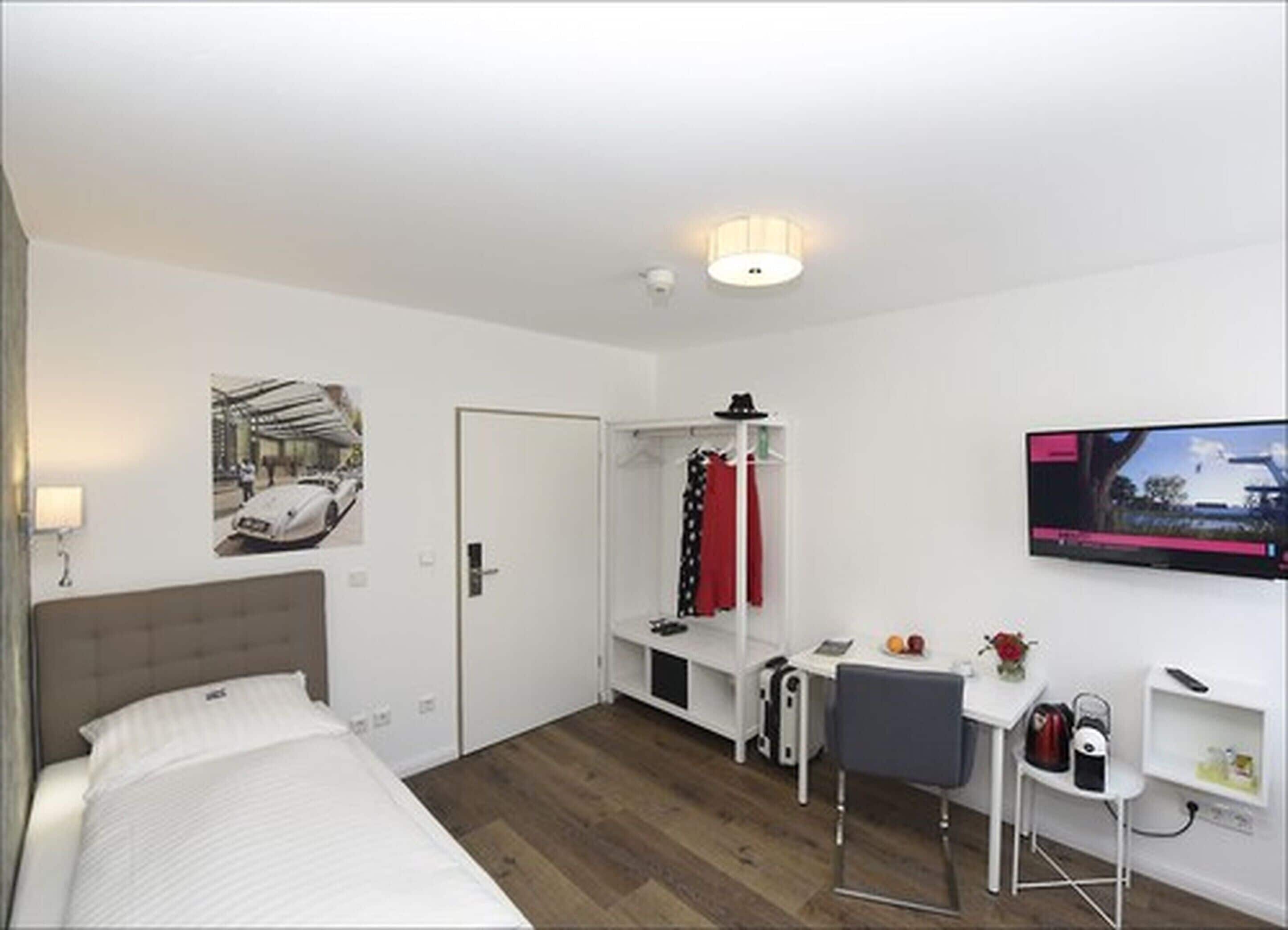 Ein Hotelzimmer mit Bett, Schreibtisch, Sessel, Fernseher, Kleiderständer und Kleiderschrank.