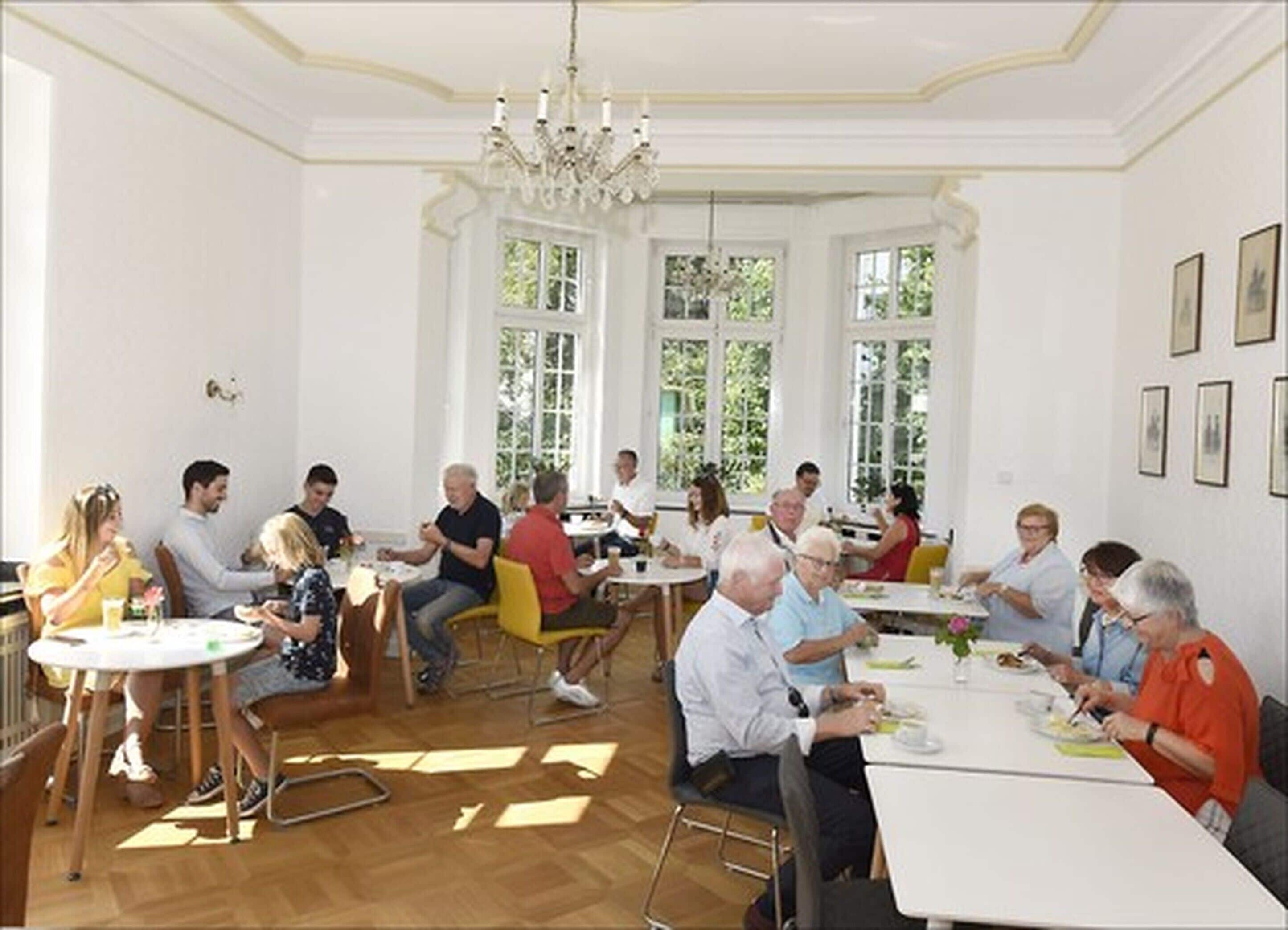 A dining area with round tables and chairs, large windows, and a chandelier.