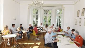 A dining area with round tables and chairs, large windows, and a chandelier.
