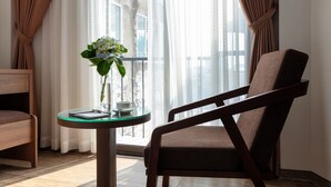 Une pièce avec une chaise en bois, une table en verre ornée d’un vase de fleurs et une vue sur l’extérieur à travers des rideaux transparents.