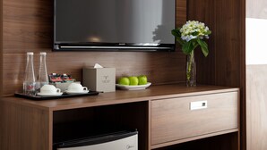 Une chambre d’hôtel équipée d’un téléviseur à écran plat fixé au mur, d’un bureau en bois sur lequel se trouve un plateau avec des articles pour le petit-déjeuner, d’un vase avec des fleurs et d’un mini-bar.