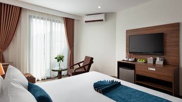 A hotel room with a bed, a desk with a flat-screen TV, a chair, a small table with a vase of flowers, and an air conditioning unit.