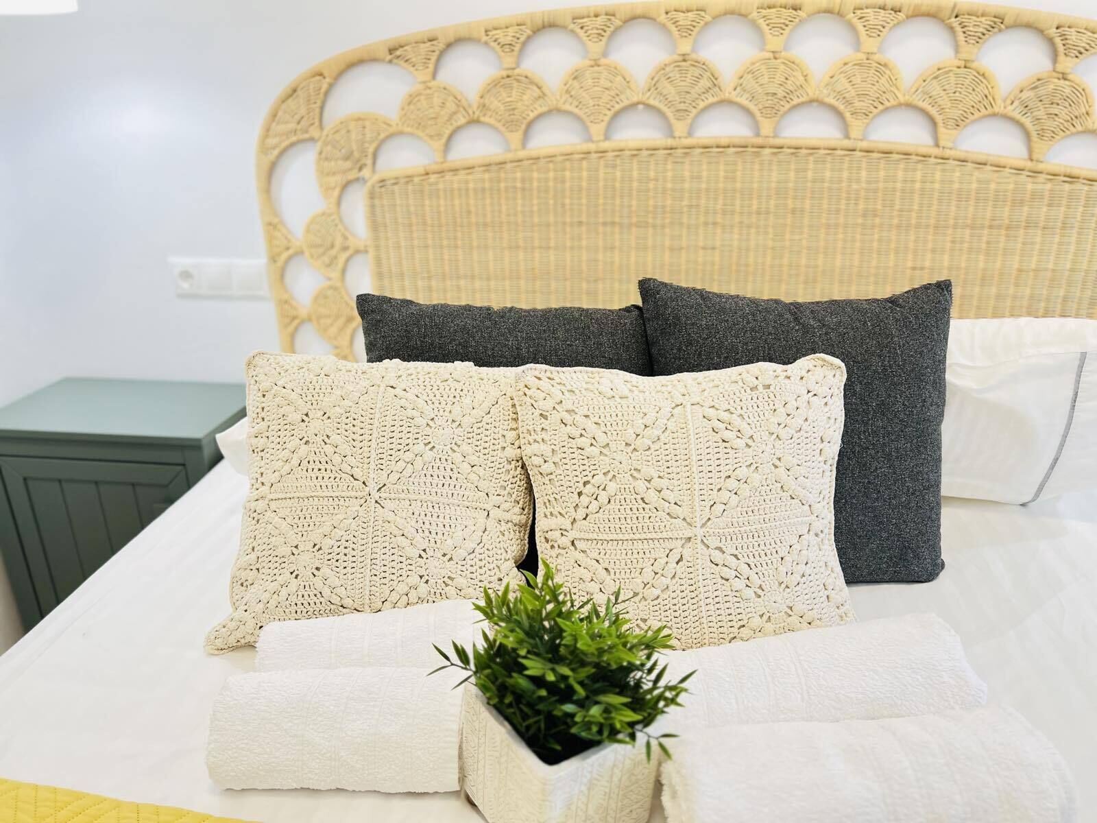 A bed with a woven headboard, two white pillows, two dark pillows, and a small potted plant.