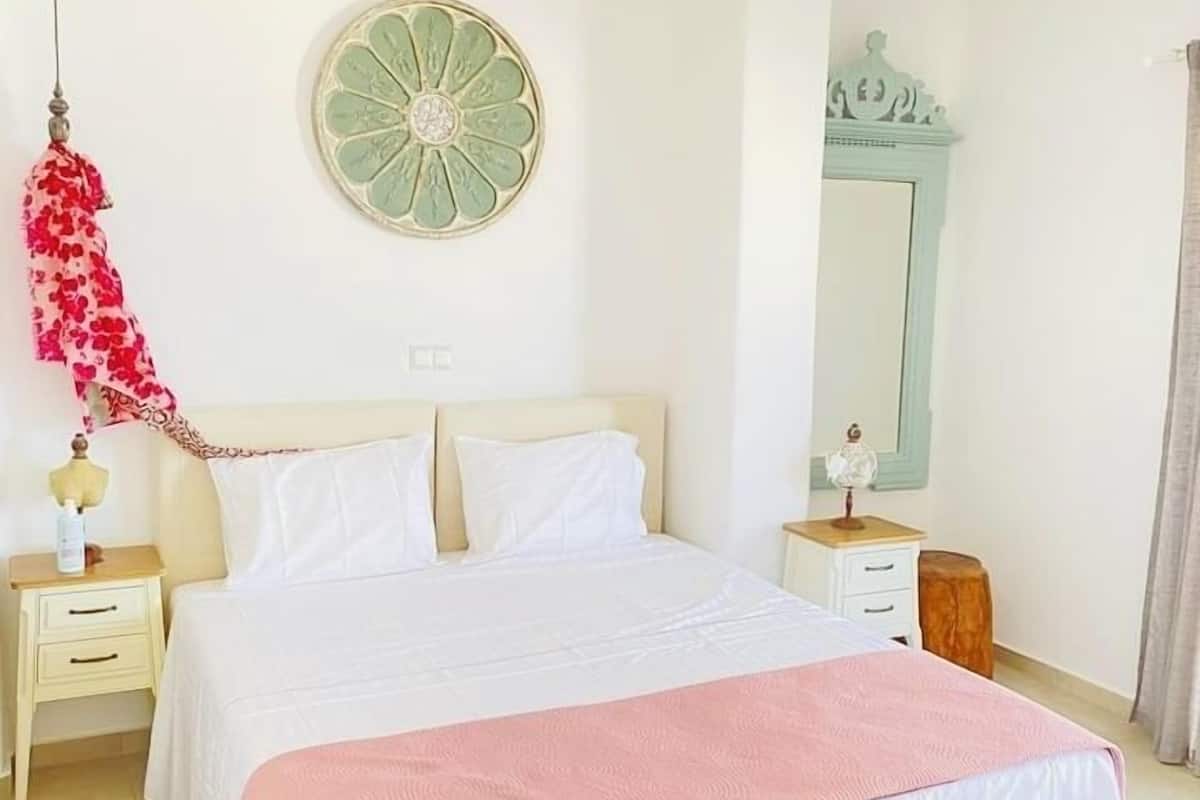 A neatly arranged bedroom with a large bed, a bedside table, a nightstand, a wall-mounted decorative item, and a mirror.