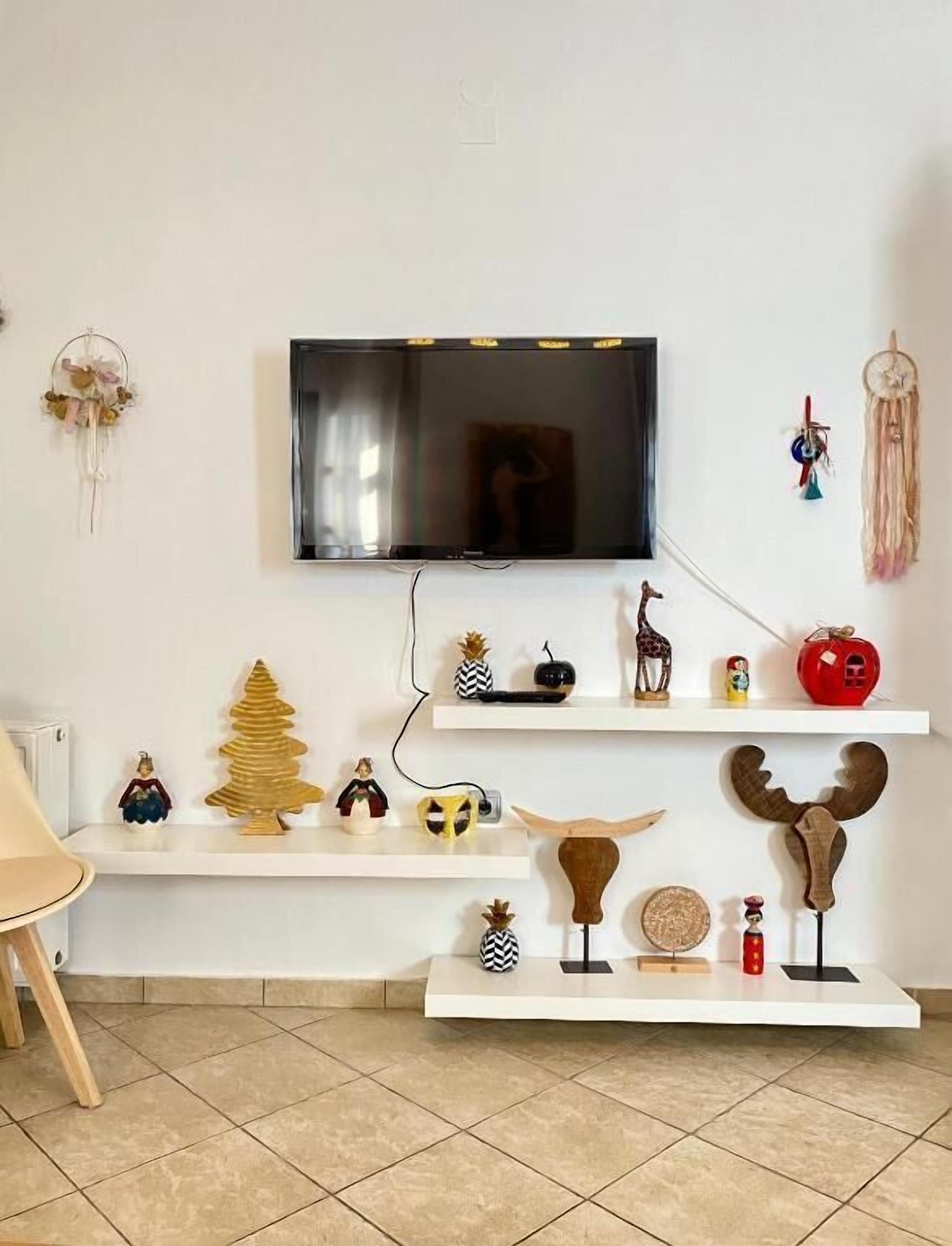 A living room with a flat-screen TV mounted on the wall, wooden shelves with various decorative items, and a dreamcatcher on the wall.