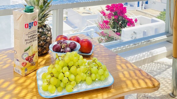 A table with a banana soursher juice carton, pineapple, grapes, plums, and an apple, with a view of the sea and white buildings in the background.