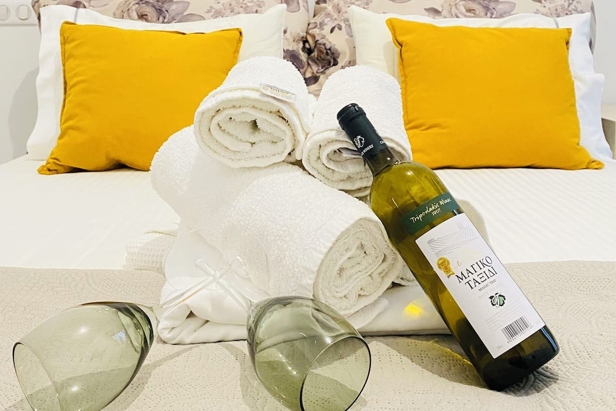 A neatly made bed with yellow pillows, rolled white towels, and a bottle of wine on a textured white surface.