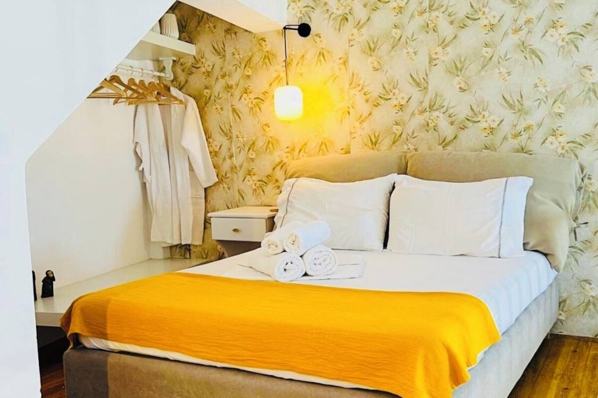 A neatly made bed with a yellow blanket, a bedside table with a lamp, and a wall with floral wallpaper.