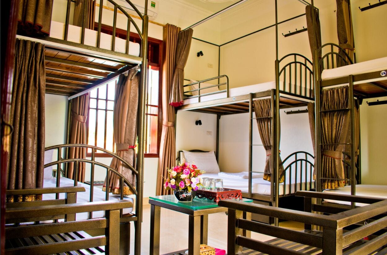 A dormitory-style room with bunk beds, a small table with a vase of flowers, and a window with curtains.