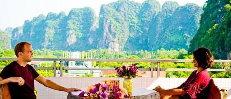 A man and a woman sitting at a wooden table with a vase of flowers, overlooking a mountainous landscape.