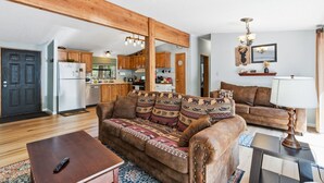 Ein Wohnzimmer mit Sofa, Couchtisch und Fernseher. Es gibt eine Küchenzeile mit Schränken und einem Kühlschrank.