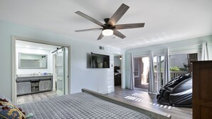 A bedroom with a bed, a ceiling fan, a TV, a bathroom with a mirror, and a sliding glass door leading to a balcony. - San Clemente / Dana Point- (San Clemente)