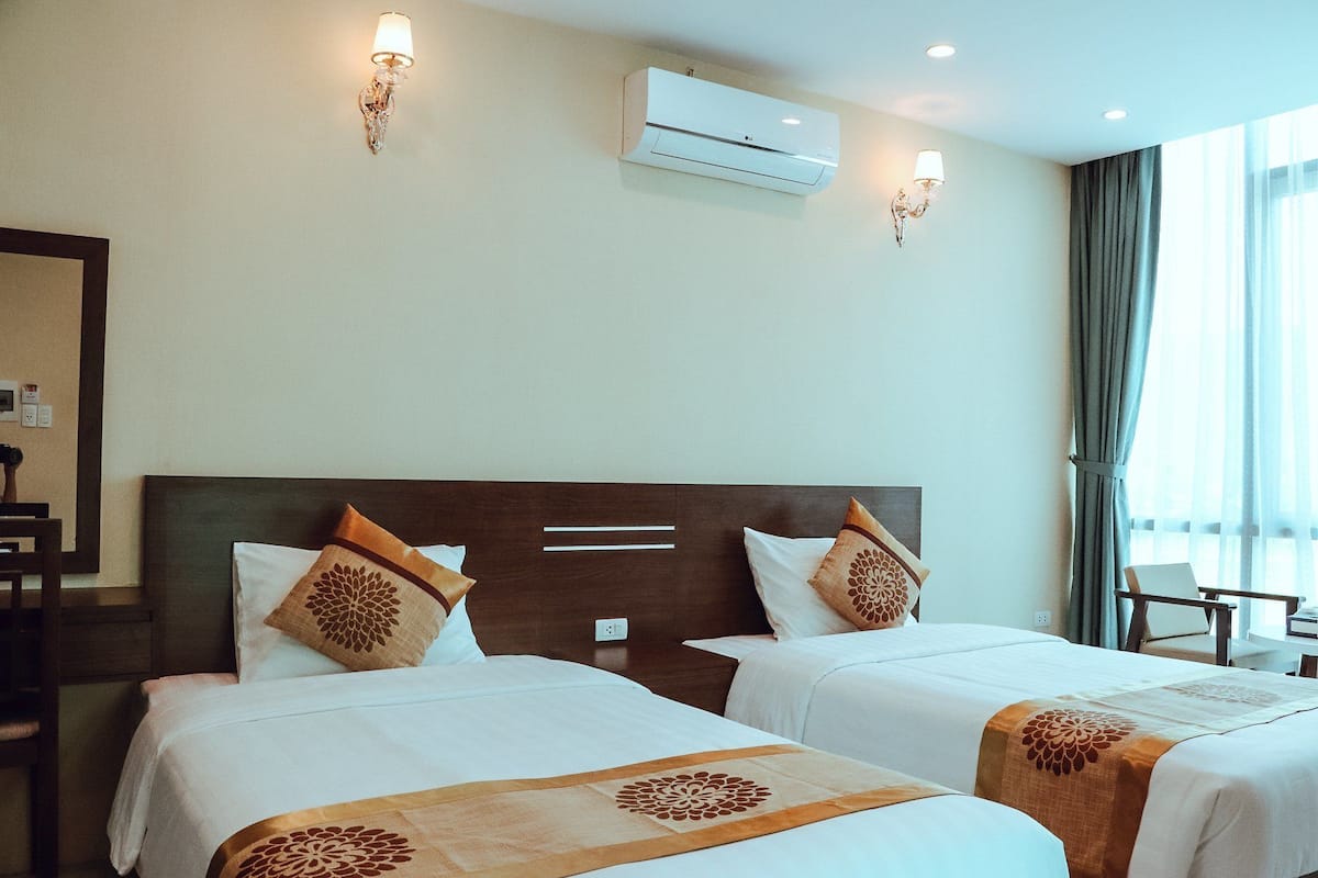 A hotel room with two beds, a large window with curtains, an air conditioning unit, and wall-mounted lamps.