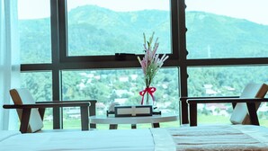 A room with a bed, two chairs, a small table with a book and a vase of flowers, and a large window with a view of green hills. - Nha hang & Khach San Tay Bac (Tuan Giao)
