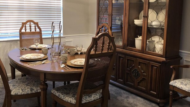 Comedor con una mesa redonda dispuesta para cuatro, rodeada de sillas de madera con asientos acolchados. Un aparador de madera con puertas de cristal está contra la pared, y un candelabro cuelga del techo.