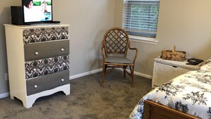 Un dormitorio con cama, una silla de madera, un aparador, televisor y ventana con persianas.