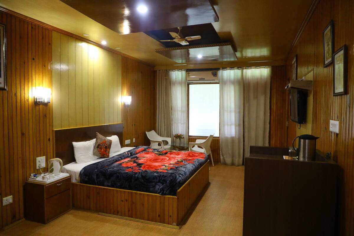 A hotel room with wooden paneling, a bed with a floral bedspread, a desk with a kettle, and a ceiling fan.