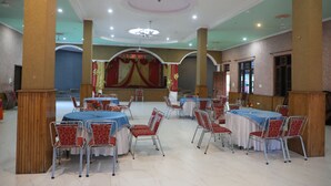 Un salón de banquetes con mesas y sillas redondas, un escenario con cortinas y una araña.